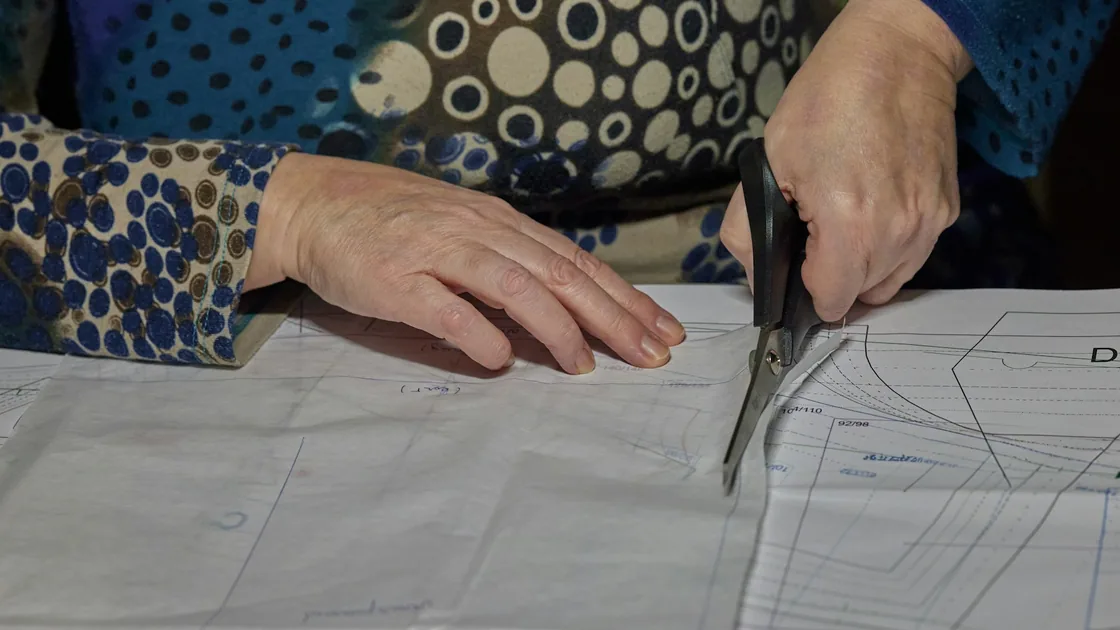 Manos recortando con tijeras una pieza de patrón de papel sobre la mesa de corte de un taller de vestuario